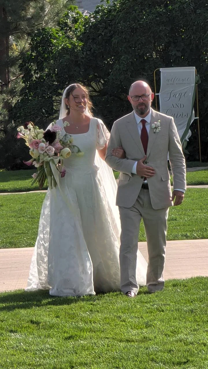 Sage walking arm in arm with a man across the lawn, holding her wedding bouquet.