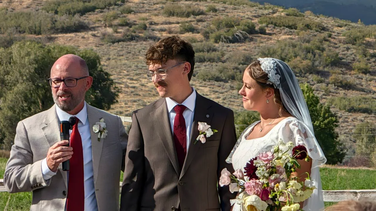 Sage and Quentin beside a man speaking into a microphone during an outdoor wedding gathering.