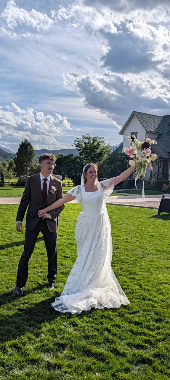 Sage raises her bouquet while holding Quentin’s hand on a sunlit lawn beneath a cloud-filled sky.